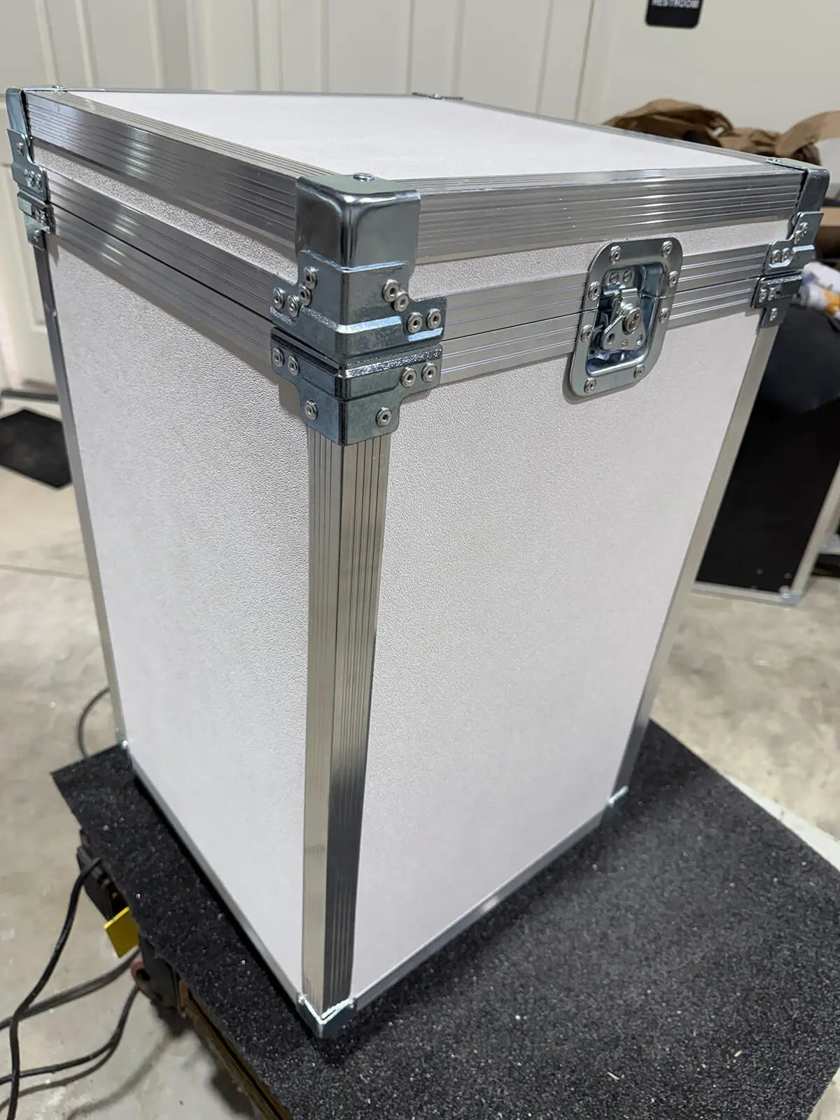 A white textured hard case with silver aluminum corner protectors, hinges, and a metal latch, sitting on a black surface in an indoor setting.