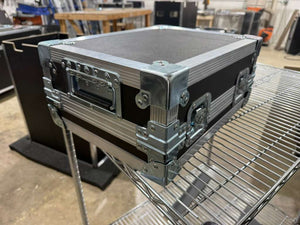 A black and silver aluminum equipment case with reinforced corners and latches, shown resting on a metal wire shelf in a workshop or studio setting.