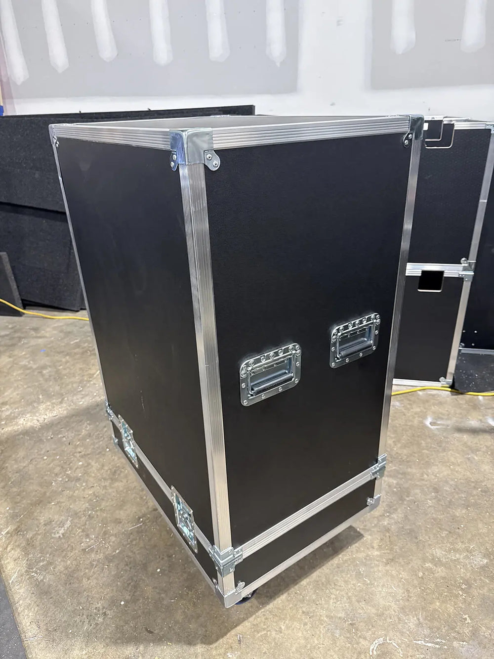 A large black equipment storage or transport case with silver metal trim, corner protectors, and two front handle grilles, photographed in a warehouse-like setting on a concrete floor.
