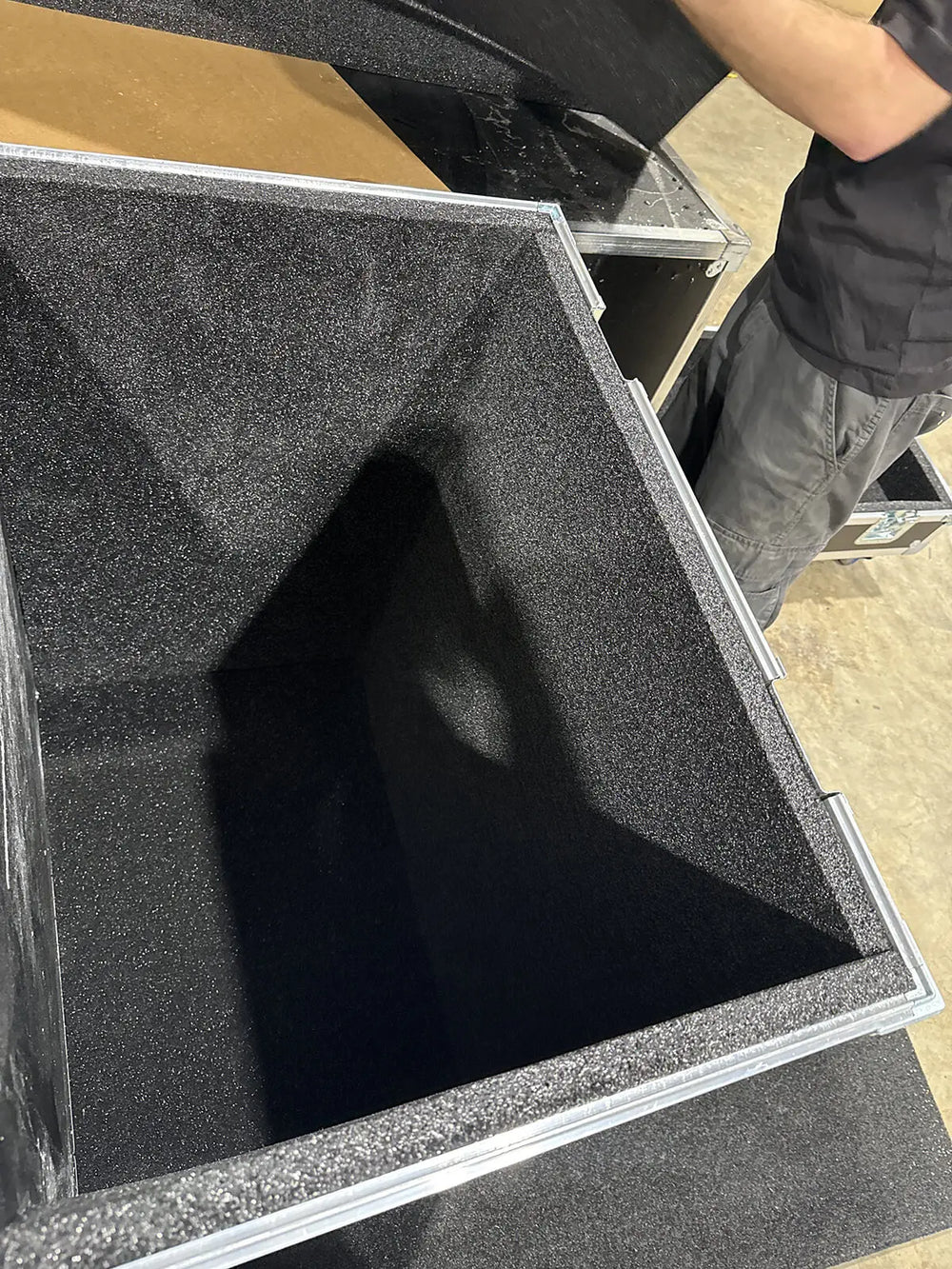 A close-up view of a large black foam-lined equipment case or storage bin with a silver metal frame, with a person's arm and torso visible in the upper right corner.