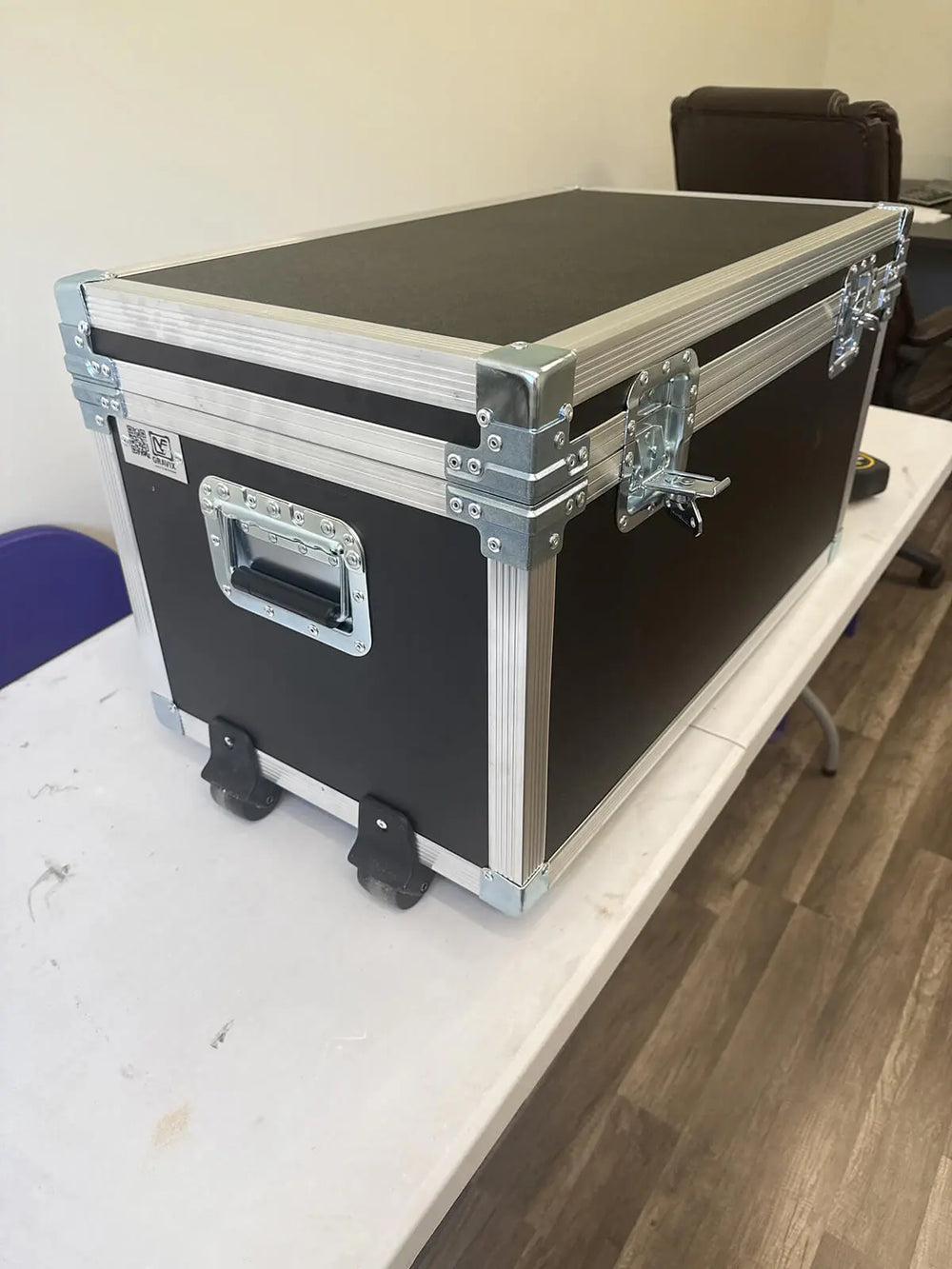 A black hard-shell equipment case with silver metal corner protectors, latches, and handle, sitting on a white table in an indoor room with wood flooring.
