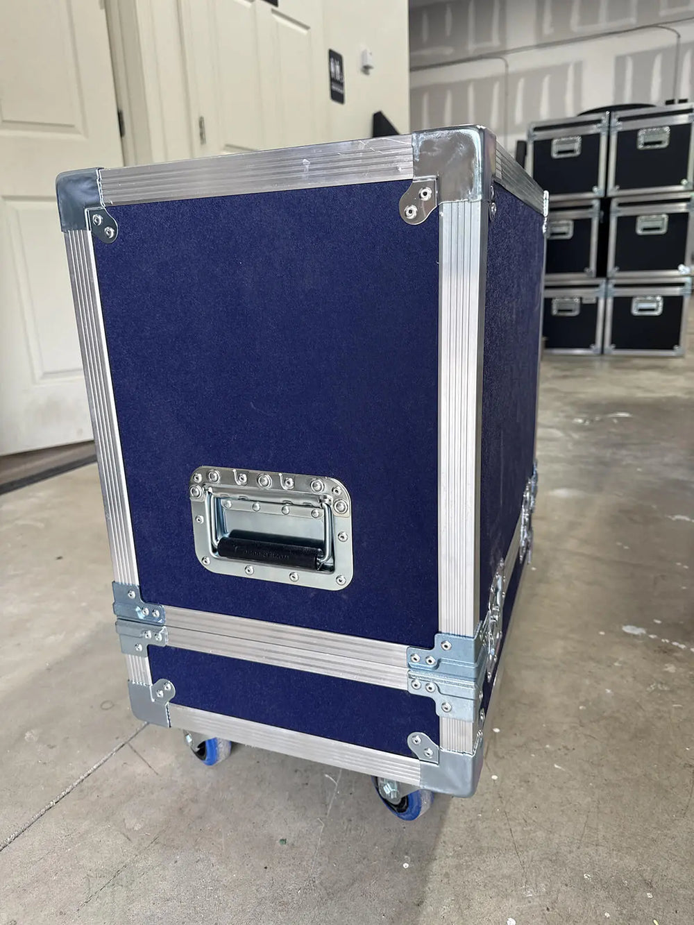 A dark blue hard case with silver metal corner protectors and a silver handle, standing on wheels in a warehouse or storage area.